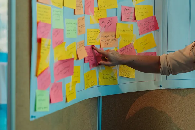 Eine Hand zeigt auf ein Projektmanagement-Board, auf dem zahlreiche farbige Post-its mit verschiedenen Beschriftungen angeordnet sind.